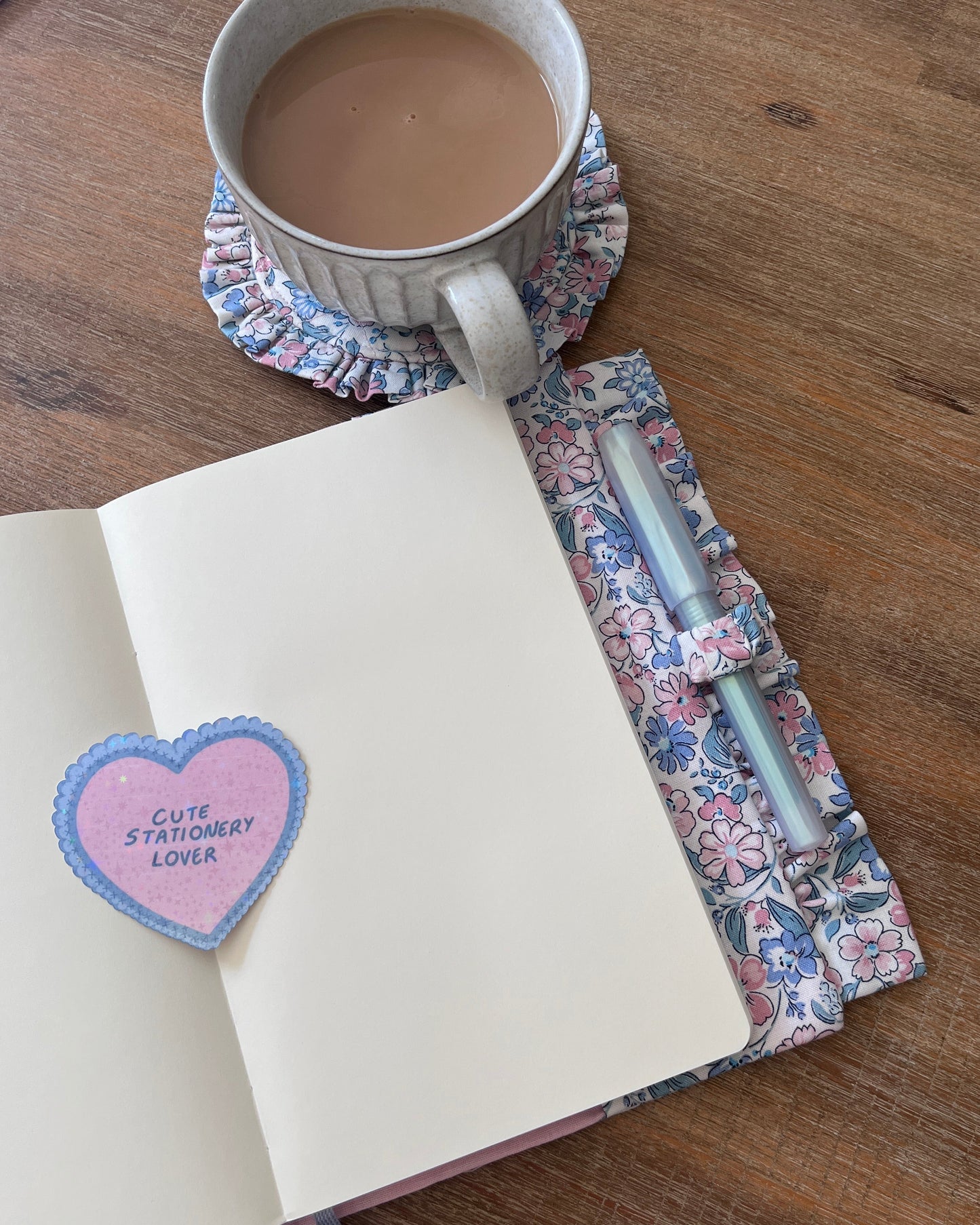 An open notebook with blank cream pages on a wooden table, featuring a pink heart-shaped sticker that reads 'Cute Stationery Lover' with a blue stitched border. A floral fabric pen loop holding two light blue pens sits on the right, and a ceramic mug filled with coffee sits on a matching floral coaster above.
