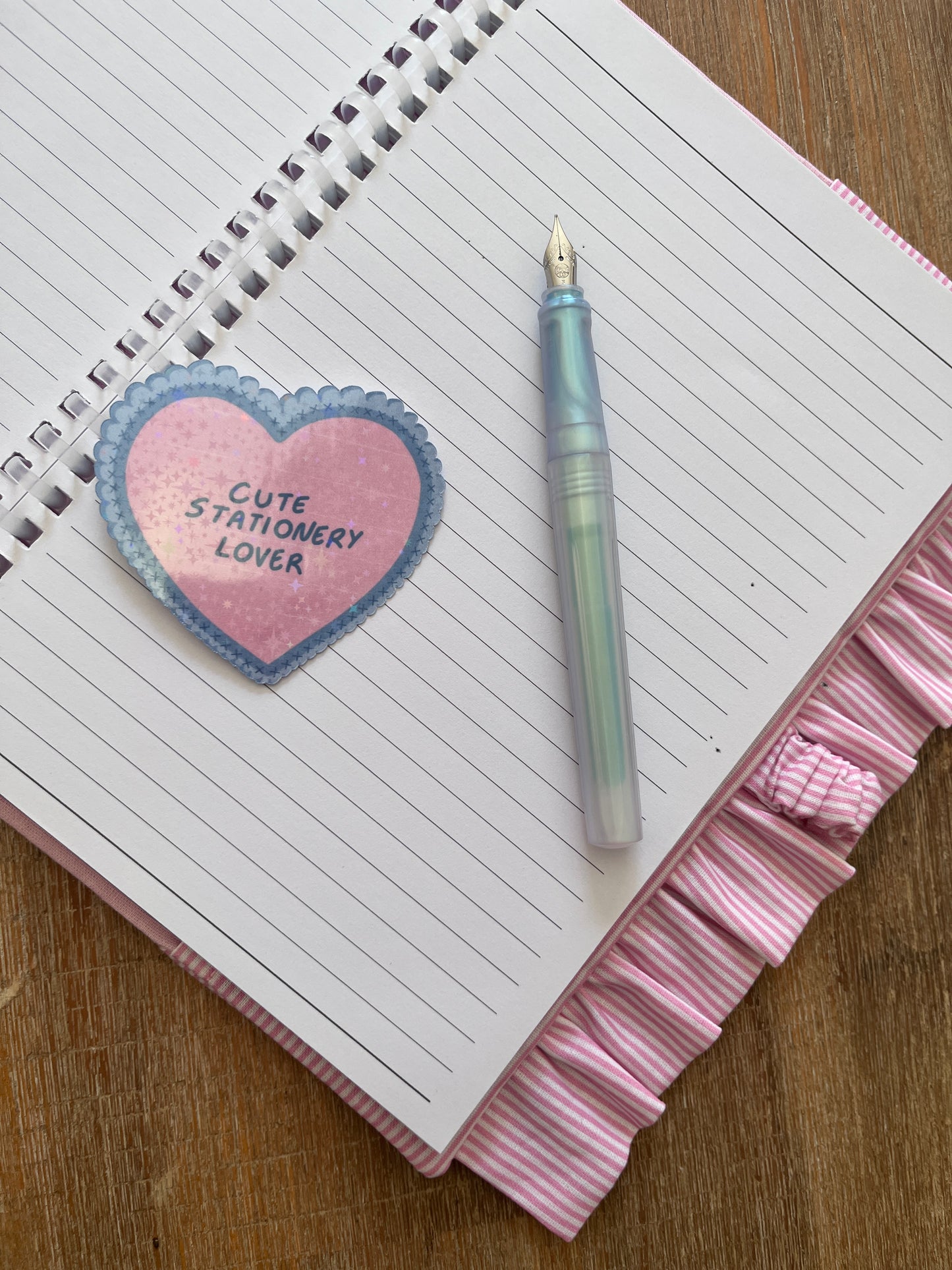 A pink and white striped spiral-bound notebook with a ruffled edge, featuring a pink heart-shaped sticker that reads 'Cute Stationery Lover' and a pastel iridescent fountain pen on a wooden surface.