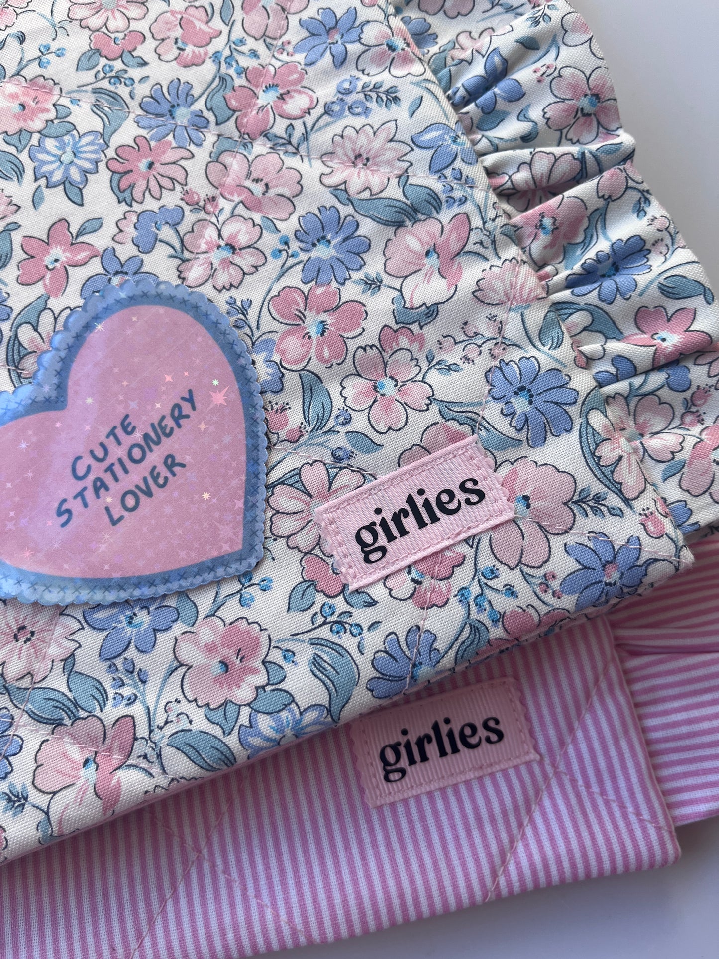 A folded floral fabric item, likely a quilt or blanket, featuring a pink and blue floral pattern on a white background, layered over a pink and white striped quilt. A pink heart-shaped patch reads 'Cute Stationery Lover' and a pink fabric tag reads 'girlies'.