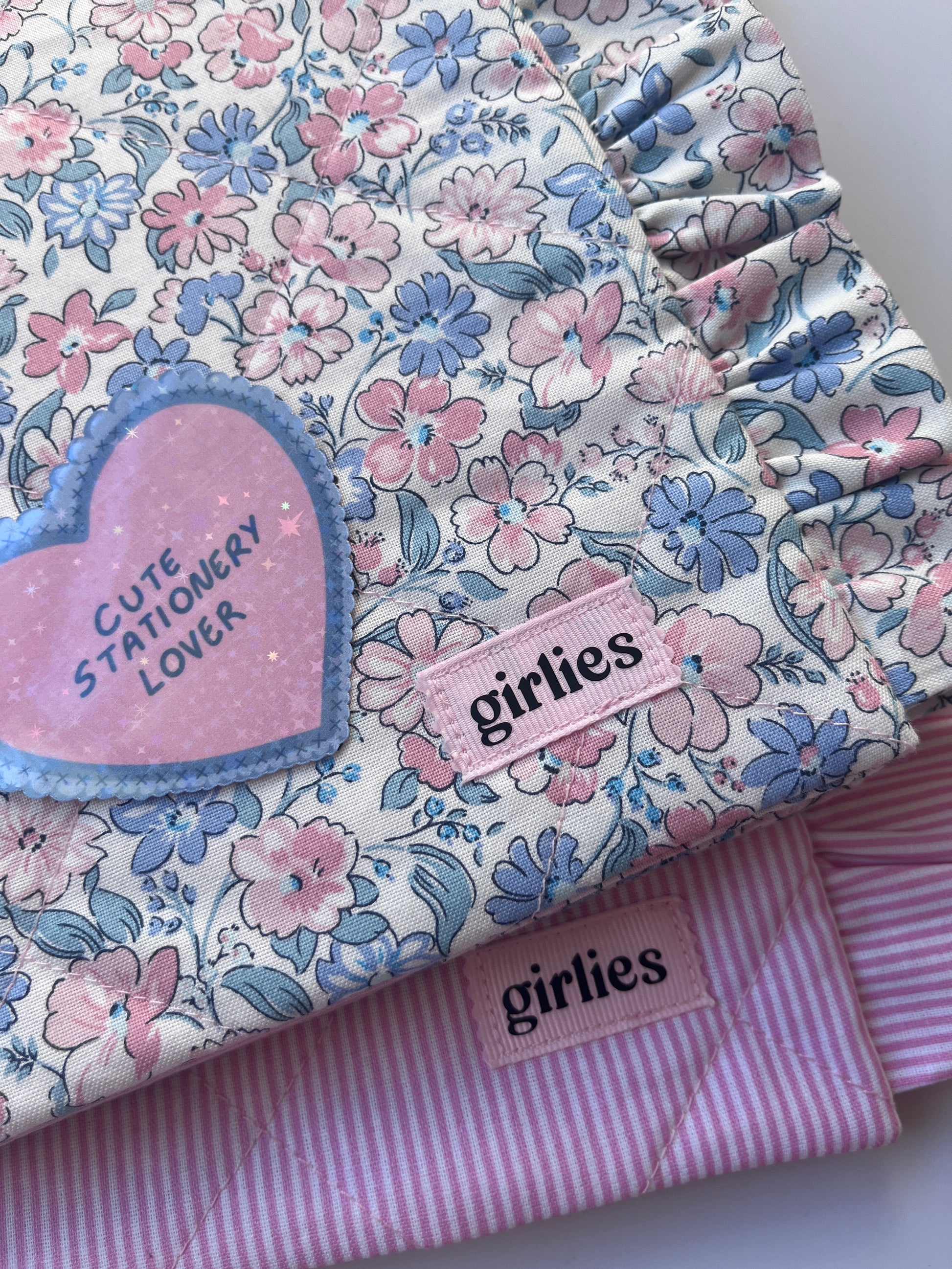 A folded floral fabric item, likely a quilt or blanket, featuring a pink and blue floral pattern on a white background, layered over a pink and white striped quilt. A pink heart-shaped patch reads 'Cute Stationery Lover' and a pink fabric tag reads 'girlies'.