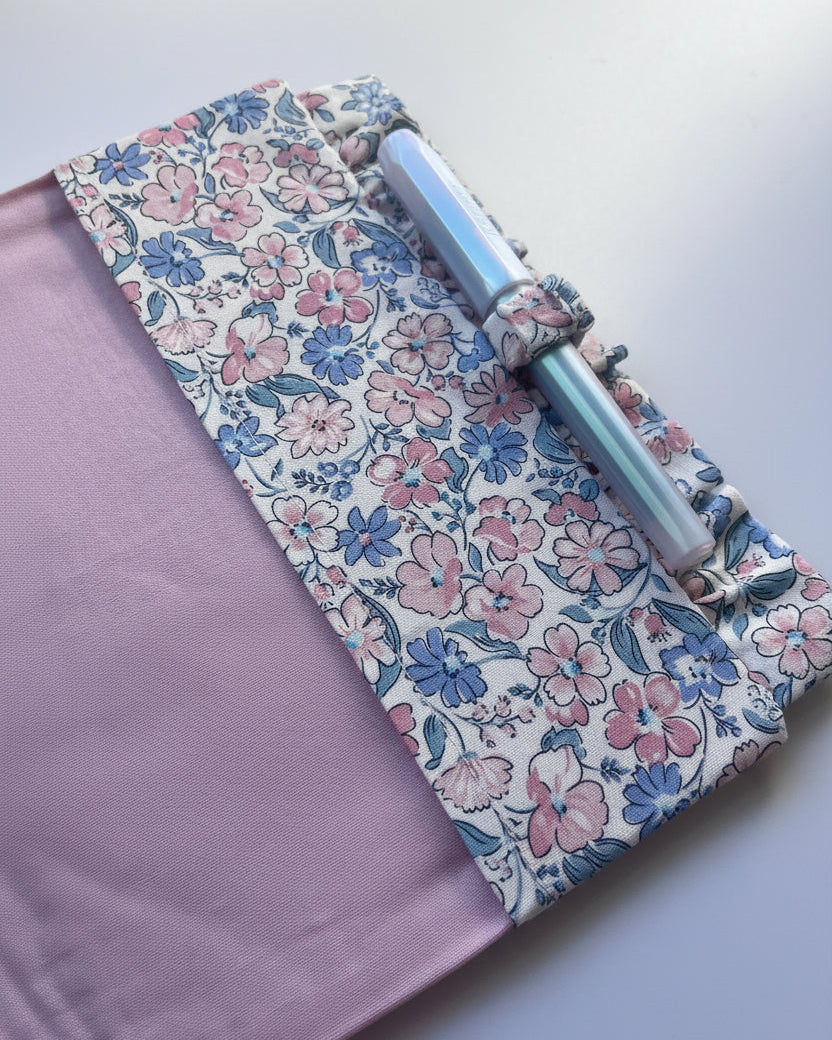 A close-up of a fabric-covered pen holder or notebook cover with a floral print on one side and a solid dusty lavender fabric on the other, holding a metallic pen.