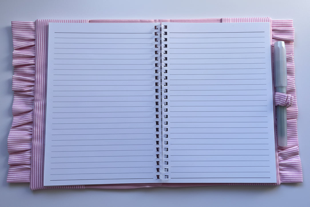 An open spiral-bound lined notebook with pink and white striped fabric covers and a matching pen loop, shown on a plain light background.