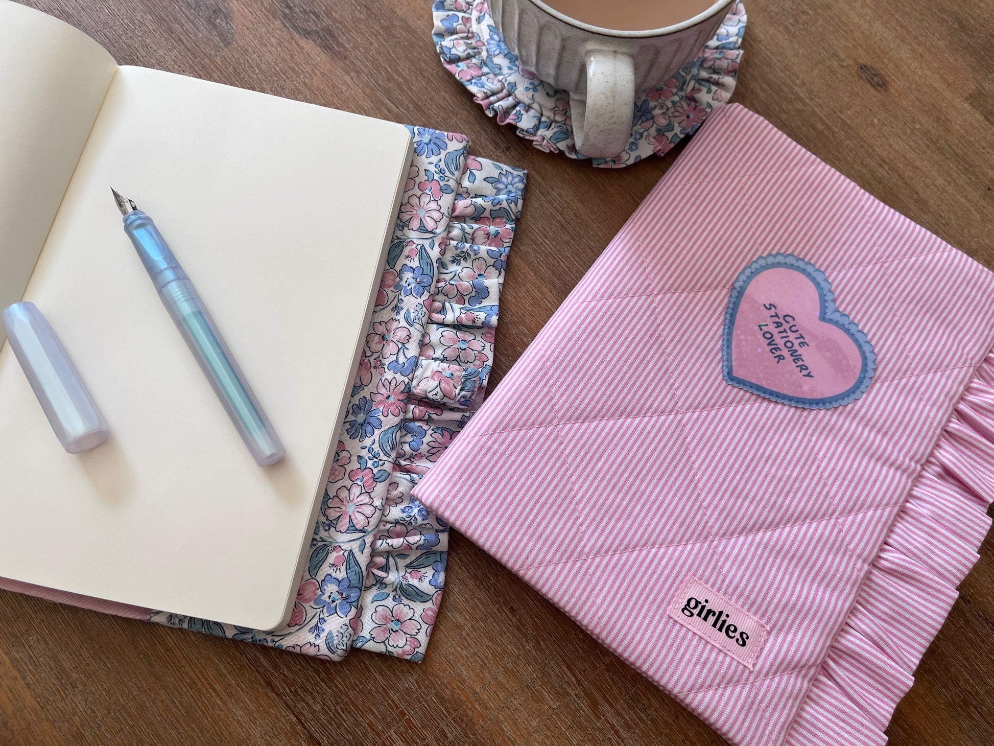 A pink quilted fabric notebook cover with ruffled edges and a heart patch, shown on a wooden table alongside an open notebook, a pen, and a mug on a coaster.