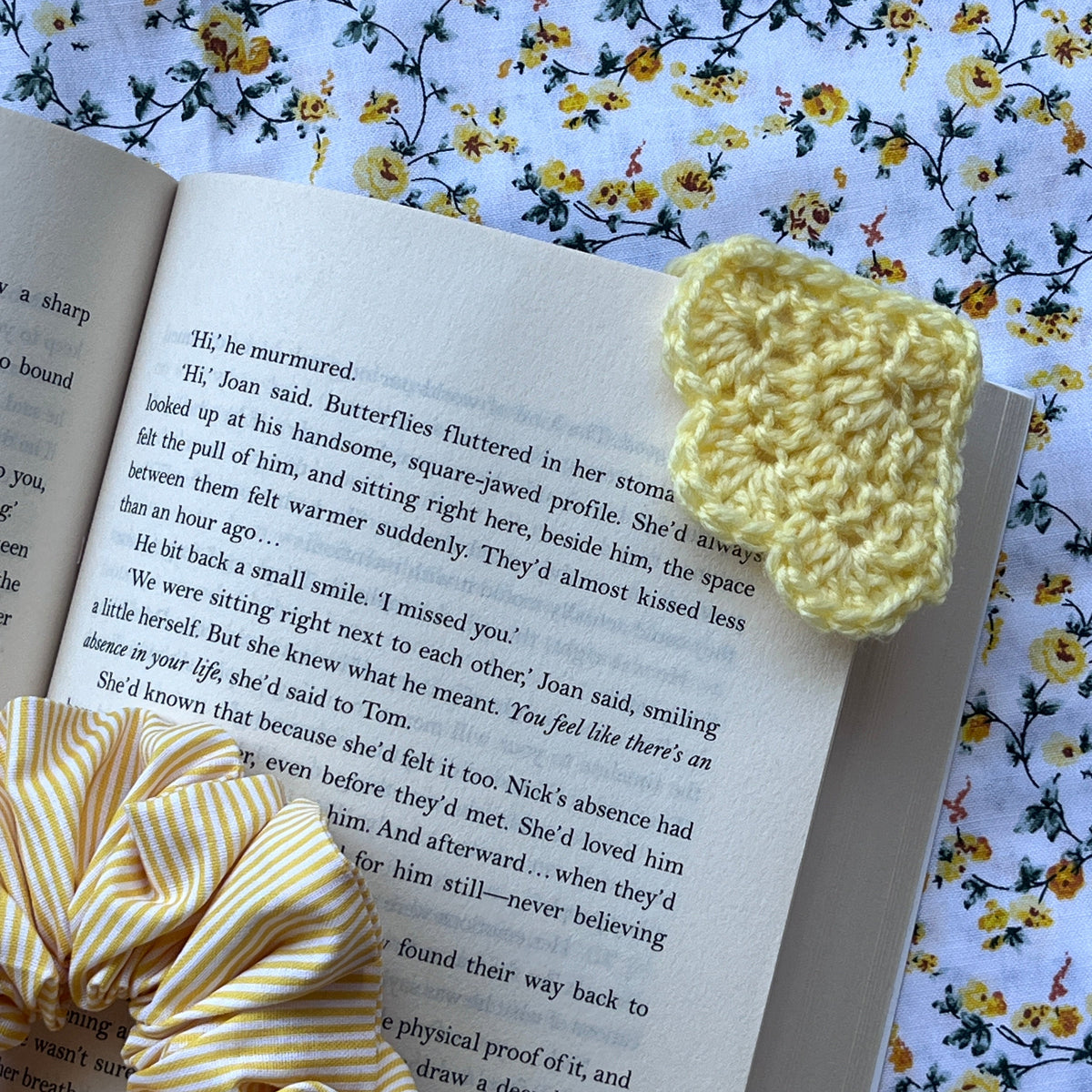 A yellow crochet bookmark rests on an open book and a floral fabric background.