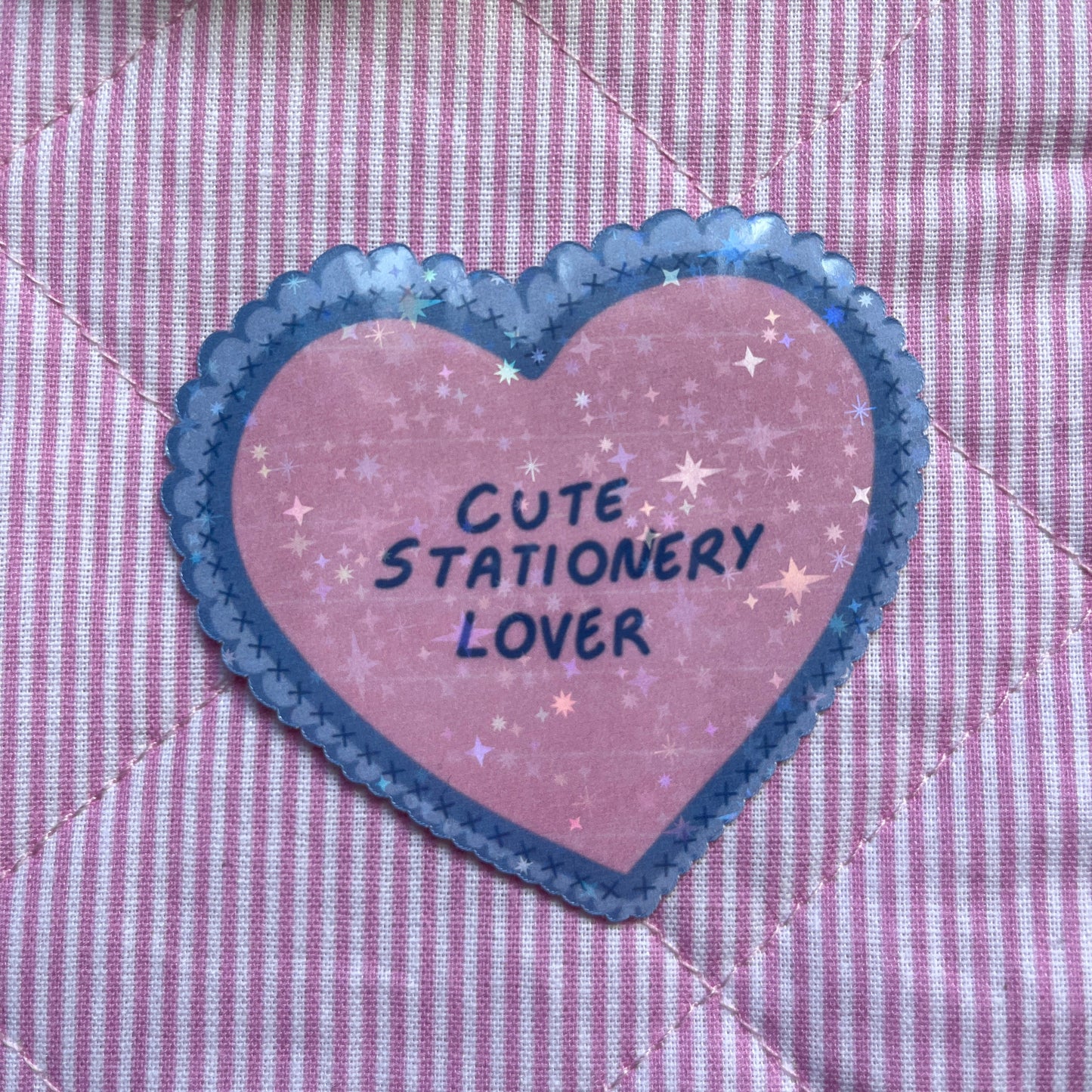 A heart-shaped sticker with a pink glittery background, blue scalloped border, and the text 'CUTE STATIONERY LOVER' in navy lettering, shown on a pink and white striped quilted fabric.