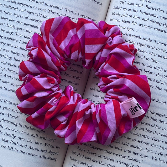 Three pink and red striped fabric hair scrunchies arranged on an open book. One scrunchie has a small pink tag